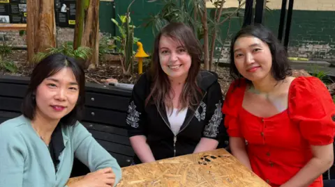 A landscape image of three women smiling at the camera. All with dark brown hair and brown eyes. The woman on the left is sitting with her arms folded and is wearing a light blue cardigan. In the middle, the woman is wearing a black hoodie and white t-shirt with her hands in her lap. Also with her hands in her lap is a woman dressed in a red dress.