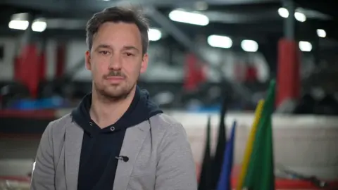 Jamie Niblock/BBC Jacques looks directly at the camera. He is beside a karting track and is wearing a grey blazer over a black hoodie. Behind him are the flags used motor racing: green, yellow, blue, black and chequered.