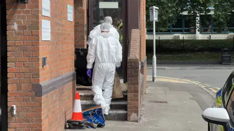 Two people wearing white forensic suits and purple gloves entering a brick building on Bedford Street. One of them is carrying a brown paper bag as he walks up the stairs into the property. A police car can be seen parked to the right.