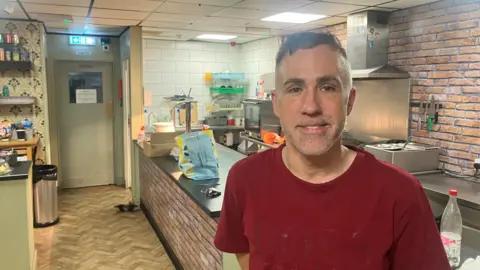 Andy Flaherty in the kitchen of his community cafe. He is wearing a red t-shirt and has short, cropped dark hair and stubble. He stands in front of a counter with a hob, grill and hot plate in the background.