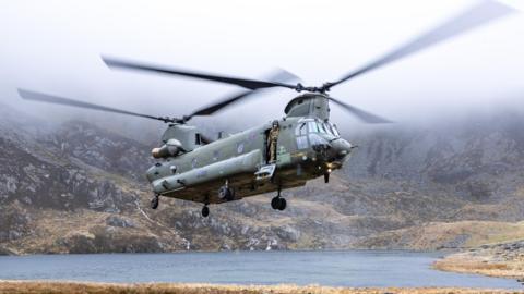 A Chinook helicopter hoovering above the ground in Eryri national park, with a crew member stood in doorway and cloud above rotor blades