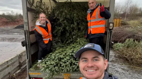 Three people at the back of a van filled with Christmas trees look to the camera, smiling. They are wearing dark clothes with orange high-vis jackets on. There are more Christmas trees on the ground