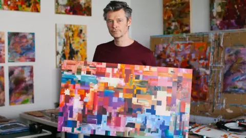 A man with grey hair in a burgundy top is standing in an art workshop with colourful abstract art in the background. He is holding a canvas in a rainbow of colours that are painted in large pixel-like squares.