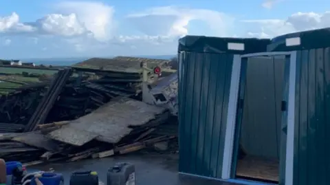 Flicka Foundation The picture shows a damaged outdoor area with storm‑hit structures. A collapsed wooden building or shelter lies in a heap on the left, with broken panels, beams and roofing scattered across the ground. On the right, a green metal structure stands partly upright with its door open, looking dented or distorted by strong winds. 
