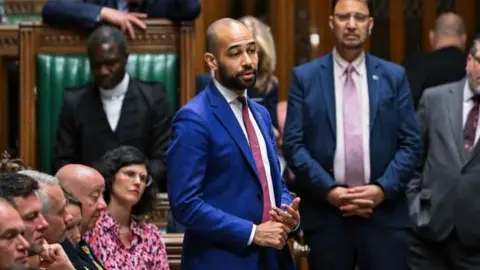 A man in a cobalt coloured suit and a red tie standing up and talking in parliament.