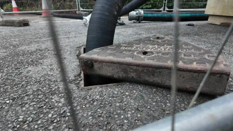 A pipe can be seen going into a hole past a lifted manhole cover inside a fenced off works compound.