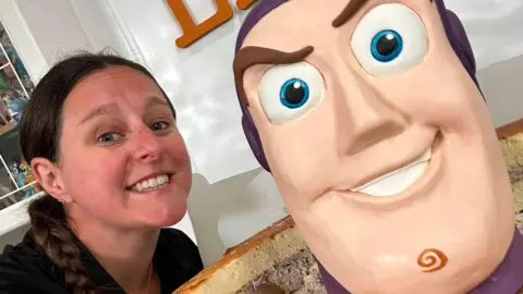 Lara Cakes A close-up photo of a woman who has long dark brown hair in a plait next to the top of a cake which has been designed like the face of Buzz Lightyear. 