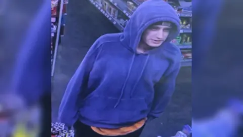 A CCTV image of a man in a shop. He is wearing a blue hooded jumper with the hood up. He has an orange t-shirt underneath it.