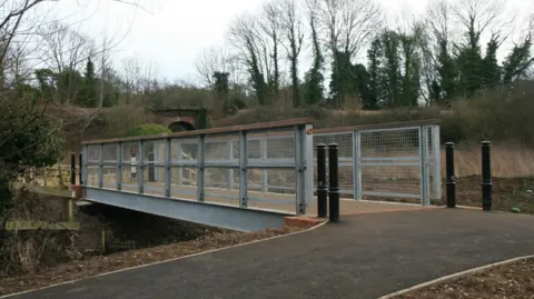 Suffolk County Council Metal and wooden bridge over a river, hills and trees in the background. A tarmac path leads up to the bridge and there are bollards on both sides