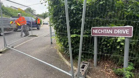 two workers in high-viz jackets inside a fenced-off area of a street with a sign saying Nechtan Drive