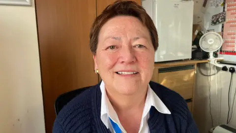 A woman wearing a navy jumper and white shirt sits in a chair. A fridge, cupboards and a fan can be seen behind her, alongside a calendar.