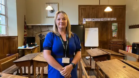 A photo of a Lorraine Curry who is the executive head of Kenton Primary School. She is standing in the middle of the photo wearing a blue quarter-length sleeved dress with stripes of black. She also has a lanyard round her neck which says staff on it. She has blonde hair past her shoulders. She is stood next to four desks made of wood. In the corner of a room there is a United Kingdom flag. 