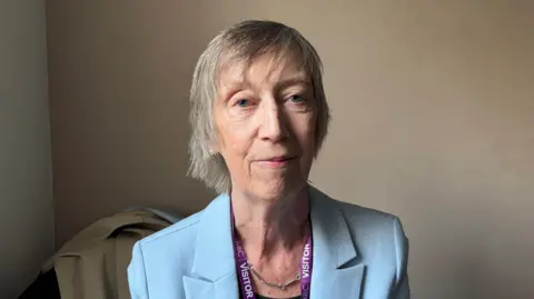A woman with short grey hair. She is looking at the camera. She is wearing a pale-blue blazer.
