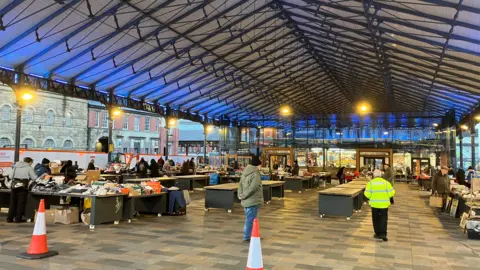 An outdoor market with a large canopy overhead. There are two orange traffic cones in the foreground, and dozens of tables stretching towards the back of the market. Traders are setting up their stalls.