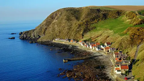 Saf37y/BBC Weather Watchers A view down to the coastal village of Crovie. The row of white-walled houses with orange roofs are at the bottom of a steep, grassy cliff. The tide is out revealing a rocky shoreline.