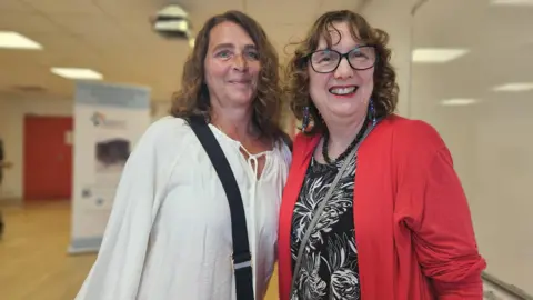 Aisha Iqbal/BBC Two middle-aged women smile into the camera. They are standing in a community hall. Both have short brown/dark blonde hair. One wears a white cotton shirt, the other wears a red jacket, thick glasses and big earrings.