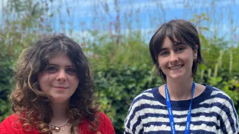 Two girls smiling at the camera. The one on the left has shoulder length curly brunette hair. She's wearing a red plattted jumper and a necklace with charms on it. The girl on the right with short black hair and earings, she's wearing a blue and white stripy jumper. 