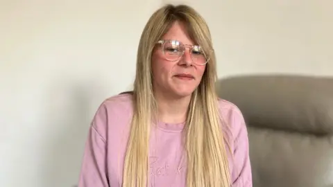 A woman with long blonde hair and glasses wearing a pink sweatshirt looking in the camera. She is sat on a sofa.