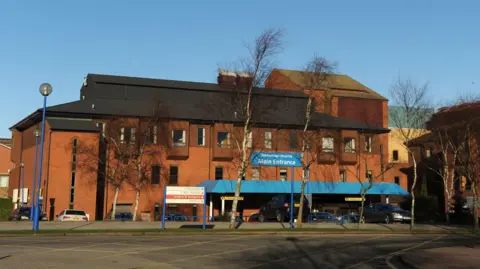 PA Media Exterior view of Scarborough Hospital’s main entrance on a clear day. The red-brick building has a modern design with multiple windows and a black roof. A blue canopy above the entrance displays the sign “Scarborough Hospital Main Entrance.”