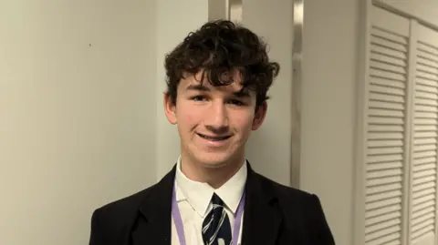 Oliver standing smiling at the camera. Behind him a white wall. He is wearing a school uniform.