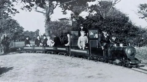 A black and white photo of a miniature railway, with a man driving a small train and several children sitting in open, trailer-style carriages in the back. Trees and a field are behind them.