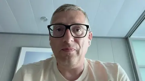 Simon Leyland has dark rimmed glasses, he is sitting in a cabin and wearing a beige tee-shirt