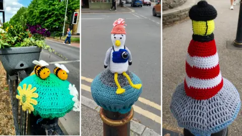 Donna Anderson Bright green crocheted bollard cover decorated with yellow flowers and two small knitted bees, next to a roadside planter and traffic lights, a crocheted seagull figure wearing a hat and carrying a blue camera, sitting on a bollard cover and a crocheted red-and-white striped lighthouse with a yellow top mounted on a grey bollard cover by a pavement.