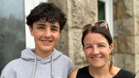 A close-up portrait photo of Jacob and his mother Amy standing shoulder to shoulder outside a stone building; one has dark curly hair, wears a grey hoodie and smiles with braces visible, while the other has dark hair tied back, sunglasses resting on their head and gold earrings. Both are looking directly at the camera, with their faces in sharp focus against a softly blurred background of stone walls and large windows.