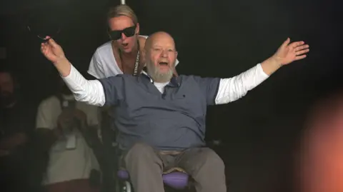 PA Media Michael Eavis is on the stage, spreading out his arms. He is wearing a T-shirt over a white long-sleeve. His daughter, Emily Eavis, is behind him pushing him onto the stage and cheering.