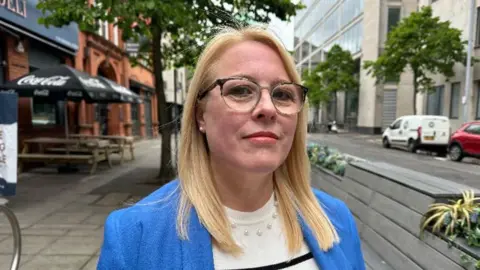 Nicola Armstrong, with blonde hair and glasses, wearing blue jacket, white and blue striped jumper and beaded necklace.