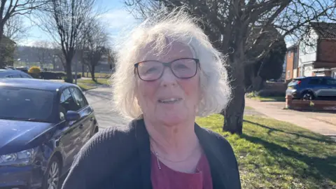 Holly Nichols / BBC A woman with grey hair wearing glasses and a black cardigan and red top standing by a tree lined road with a car in the background. 