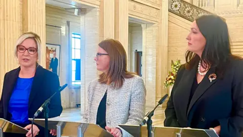 BBC From left to right of the image, Michelle O'Neill, Caoimhe Archibald and Emma Little-Pengelly stand in front of mics inside Stormont
