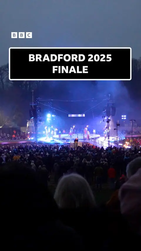 View of a brightly lit stage across the top of a crowd on a dark evening