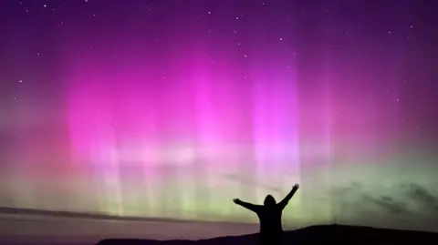 Jane Taylor The Aurora Borealis casts a purple and green shade in the sky. A figure silhouetted in shadow has their arms in the air watching it, clearly excited.   