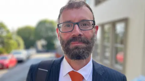 Deputy Aidan Matthews - A man with scruffy brown hair, wearing glasses. He has a bushy brown and grey beard. He is wearing a blue and white pinstriupe suit with a black backpack strap over his shoulder. 