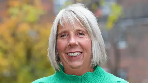 CRUK Alison Tatum dans un pull vert debout à l'extérieur avec des arbres et des bâtiments flous en arrière-plan.