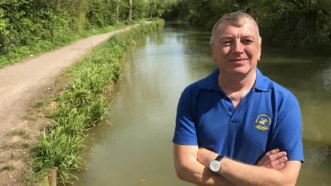 Justin Guy A man - Chair of the Melksham, Chippenham and Calne branch of the Wilts & Berks Canal Trust, Howard Wilson - with short grey hair and arms folded and wearing a royal blue polo shirt standing in front of the Pewsham canal in Chippenham. 