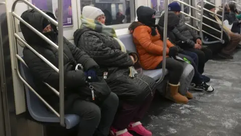 People sit bundled up with hats, scarfs and boots on a New York train