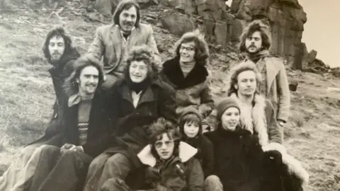 Courtesy of Geoff Bowen A black and white picture of a group pf people. They wear clothing which suggests the picture is from the 1970s. They are a mix of young men and women, with a couple of children also with them.