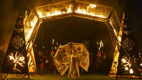 The May Queen wearing a white dress walks through a gate surrounded by fire at the start of the Beltane festival.