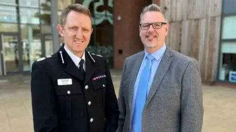 A man with grey hair, glasses and a dark suit and blue tie standing  beside a man with dark hair and a black police uniform
