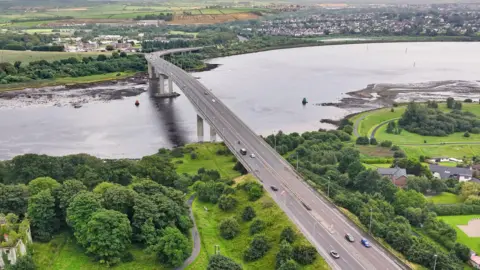 An aerial view of Foyle Bridge. A large grey bridge between two grassy land masses. A number of cars are visible crossing the bridge over the River Foyle. 