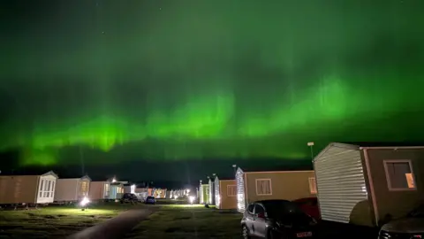 Micah Campbell Bright green aurora borealis illuminates the night sky above a row of holiday homes and parked cars, casting a vivid glow across the horizon.