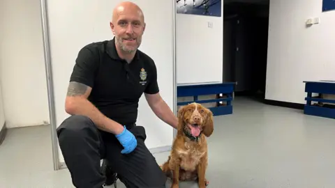 Police dog trainer, PC Paul Cooper crouched down in his uniform with blue latex gloves, next to three-year-old red cocker spaniel Baxter, who is sat with his tongue out.