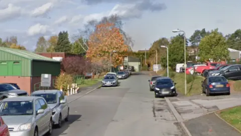 A street with cars parked on both sides with some grassy verges. Industrial-looking buildings are on both sides of the street.
