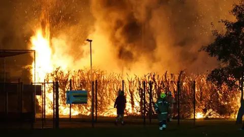 Large flames coming from hedges and trees surrounding football pitches. The sky is dark and firefighters and members of the public are standing nearby.