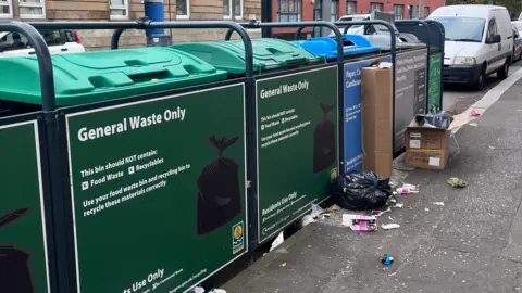 Cardboard boxes, a bin bag and litter are strewn over the ground in front of a bin hub.