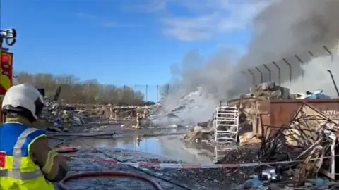 A large cloud of smoke coming off some debris while firefighters attempt to stop it with water. 