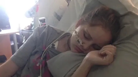 Regan Sparks A young girl is asleep in a hospital bed, with a tube in her nose. Her skin is pale and she is wearing a grey t-shirt.