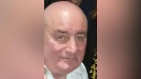 A older man looking at the camera. He is bald and wearing a white shirt